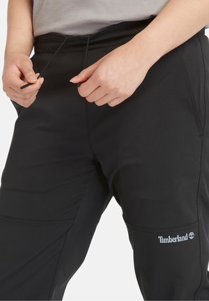 Zwarte sportbroek met een elastische tailleband en trekkoord, voorzien van zijzakken en een Timberland-logo op het bovenbeen. Soepele textuur.