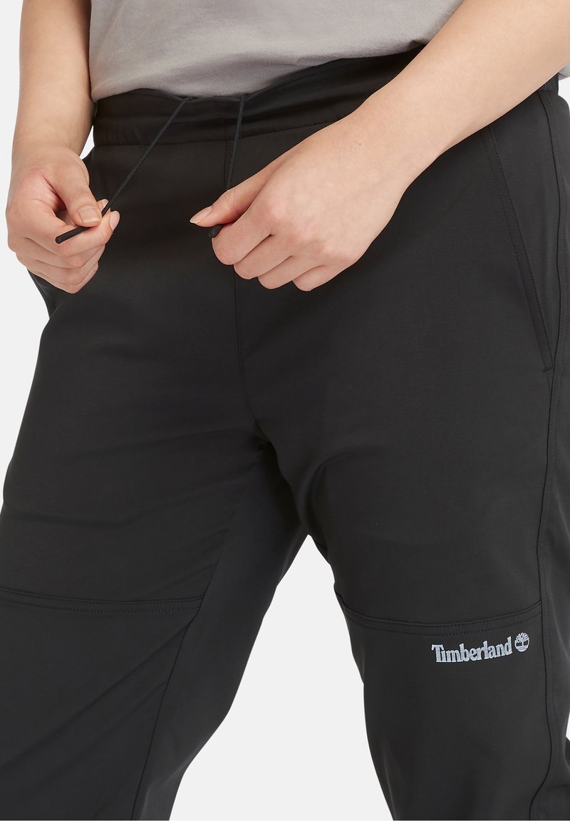 Black athletic pants with an elastic waistband and drawstring, featuring side pockets and a Timberland logo on the thigh. Smooth texture.
