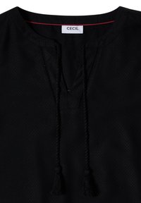 Blouse noire texturée avec une encolure ronde et deux liens à pompons tressés suspendus à l'avant, avec une étiquette blanche comportant "CECIL" à l’intérieur.