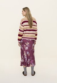 Gestrickter Pullover mit horizontalen Streifen in Creme, Burgunderrot und Pink, kombiniert mit einem langen lila Blumenkleid und grauen Socken.