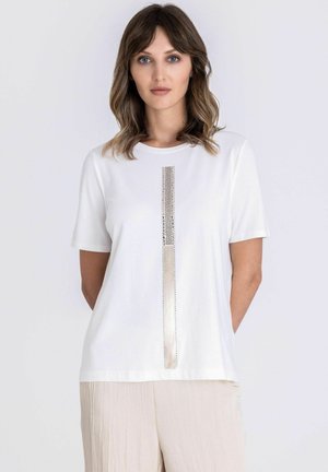 Vrouw met schouderlang bruin haar, gekleed in een wit T-shirt met korte mouwen en een verticale metalen streep, en beige broek, tegen een effen achtergrond.