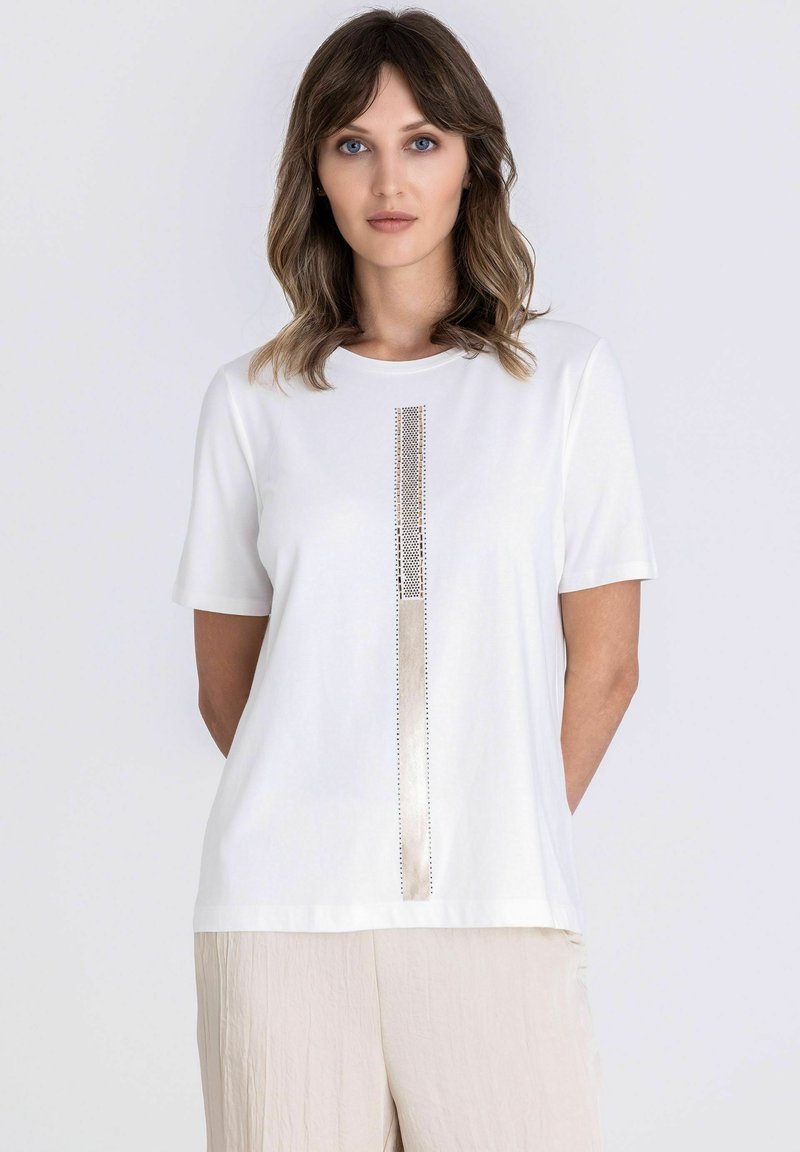 Vrouw met schouderlang bruin haar, gekleed in een wit T-shirt met korte mouwen en een verticale metalen streep, en beige broek, tegen een effen achtergrond.