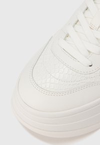 Λευκό αθλητικό παπούτσι με υφή σε σχήμα καπιτονέ στο μπροστινό μέρος, λείο δέρμα, επίπεδα κορδόνια και παχύ, μαλακό πέλμα.