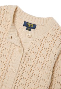 Helle cremefarbene gestrickte Strickjacke mit einem Lochmuster, gerippten Kanten und zwei Knöpfen. Das Etikett trägt "OVS kids" in Blau und Gelb.