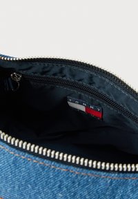 Traper torbica s tamnoplavim podstavom, srebrnim patentnim zatvaračem i bež prošivom. Unutra se nalazi etiketa marke s crvenim, bijelim i plavim detaljima.