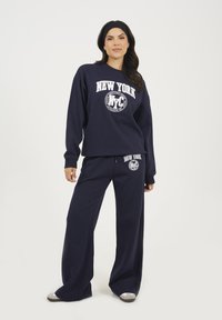 Donna che indossa una felpa navy blu con la scritta New York NYC e pantaloni larghi coordinati, in piedi su uno sfondo chiaro e semplice.