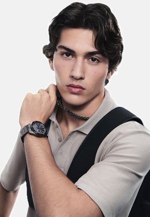 Reloj de pulsera negro con esfera redonda y correa texturizada, llevado sobre una camisa polo de color beige claro. El fondo es blanco liso.