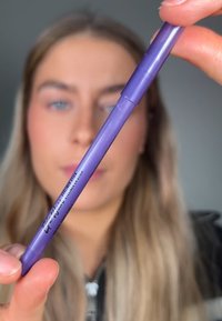 Crayon eyeliner violet avec une finition lisse et brillante. La marque est visible sur le côté. Le crayon est tenu entre les doigts.