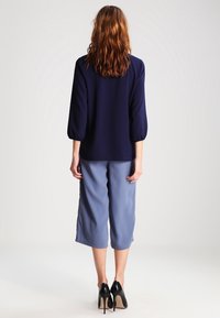 Blouse marine à manches bouffantes associée à un pantalon culotte bleu clair. Des escarpins noirs complètent la tenue, mettant en valeur une texture de tissu lisse.