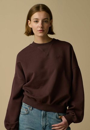 Jeune femme aux cheveux courts bruns portant un sweat-shirt marron foncé avec le mot "AMOUR" brodé, et un jean bleu clair, debout devant un fond beige.