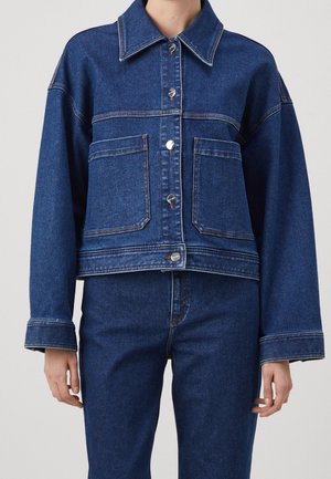 Personne portant une veste en jean bleu foncé avec des boutons argentés et un jean en denim taille haute assorti, debout devant un fond uni.