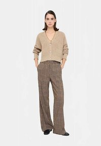 Cardigan court beige en tricot avec col en V et fermeture à boutons, associé à un pantalon large à carreaux marron, offrant une coupe décontractée.