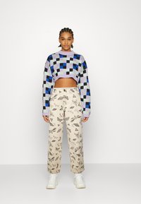 Sweater cropped de crochet em padrão xadrez roxo, azul e verde. Calças bege com estampas de insectos e folhas pretas. Ténis brancos.
