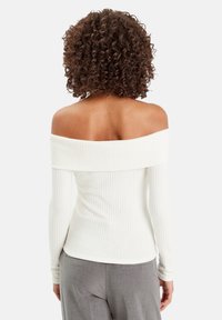 Donna con capelli ricci e scuri, che indossa un top bianco a spalle scoperte e a maniche lunghe, e pantaloni grigi, vista da dietro su uno sfondo bianco.