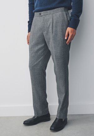 Mand iført grå plisserede bukser, sorte lædersko og en mørkeblå sweater, der står op ad en hvid væg.