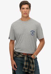 Camiseta de algodón gris con un gráfico azul en el lado izquierdo del pecho, combinada con una camisa de cuadros en tonos azul marino y verde atada a la cintura, llevada con jeans.