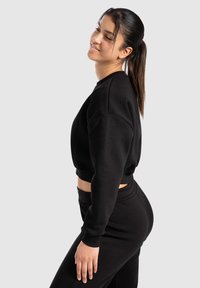 Sweatshirt noir court avec des manches longues, fabriqué en tissu doux. Présente des poignets côtelés et une coupe décontractée. Associé à un pantalon de jogging noir.