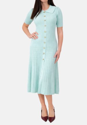 Vrouw draagt een lichtblauwe, geribbelde gebreide midi-jurk met korte mouwen, een Peter Pan-kraag, gouden knopen aan de voorkant en bordeauxrode puntige hakken.