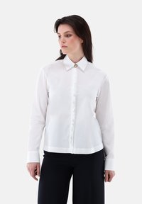 Camicia bianca con bottoni, realizzata in tessuto liscio, con colletto classico, bottoni frontali e un dettaglio decorativo sul colletto.