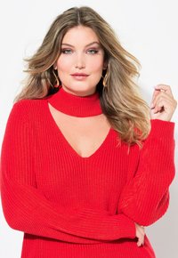 Pull en tricot rouge côtelé avec un décolleté en V plongeant et un col haut. Doté de manches longues avec de larges poignets et une finition texturée.