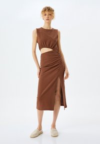 Abito marrone con tagli strategici, tessuto testurizzato, design senza maniche, arricciature laterali e spacco alto alla coscia. Modella che indossa scarpe neutre.