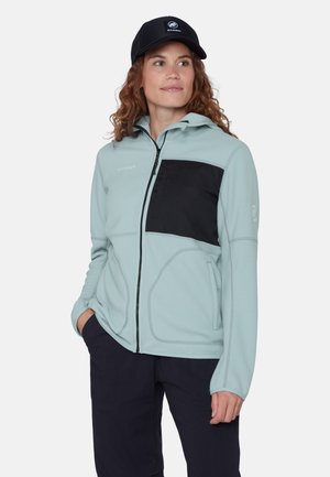 Kobieta w jasnoniebieskiej polarze zapinanej na zamek z czarną kieszenią na piersi i czarną czapką, ręce w kieszeniach granatowych spodni, patrzy w prawo.