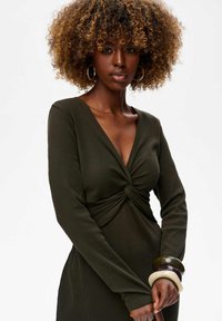 Zielono-oliwkowa sukienka z długim rękawem, z charakterystycznym skręconym węzłem na biodrach. Gładka tekstura materiału, dopasowany krój, dopełniony zestawem bransoletek.