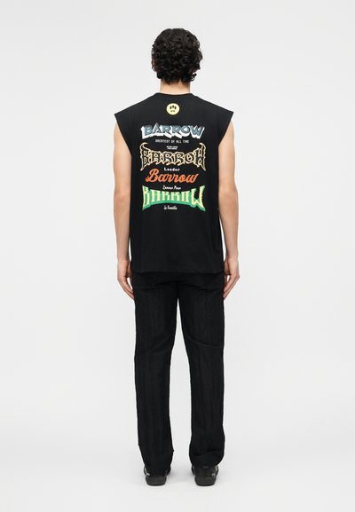 Άτομο με σγουρά μαλλιά, φοράει μαύρο αμάνικο πουκάμισο με πολύχρωμο διακοσμητικό κείμενο «Barrow» και μαύρο παντελόνι, στέκεται με την πλάτη προς τα εμπρός.