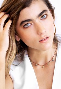 Gullringe, et flerfarget perlekjede, myk hud og lysbrunt hår. Emnet har på seg en hvit topp med dyp utringning.