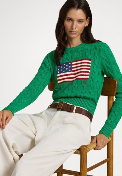 Camisola verde de malha com um desenho tricotado da bandeira americana no peito; usada com calças creme e um cinto castanho.