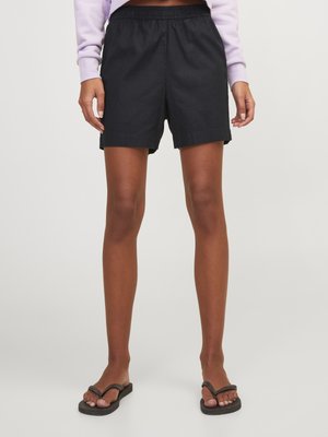 Personne portant un short noir à taille élastique, un haut à manches longues violet clair et des sandales noires, debout sur un fond clair uni.