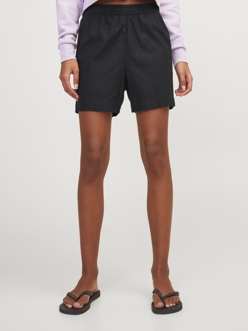 Persona que lleva puestos unos pantalones cortos negros con cintura elástica, una camiseta de manga larga de color lila claro y sandalias negras de playa, de pie sobre un fondo claro y liso.