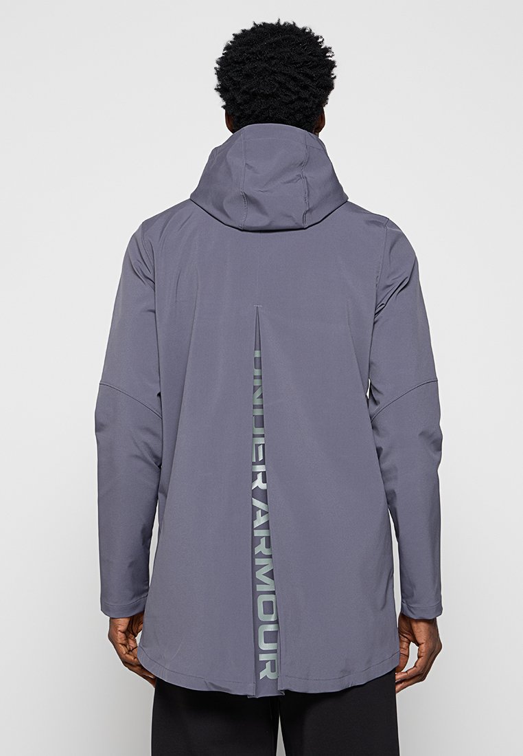 Veste imperméable grise avec capuche, présentant un dos long et une fente centrale. Texte de marque dans une couleur contrastante le long de la couture arrière.