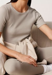 Donna seduta a gambe incrociate su un divano beige, che indossa una maglietta corta a coste color taupe e leggings a coste abbinati, con calze bianche.