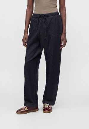 Person trägt locker sitzende schwarze Hose mit Kordelzug in der Taille, beige Tanktop und burgunderrote Sneakers mit beigen Details auf hellem Hintergrund.