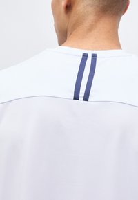 Άτομο που φαίνεται από πίσω φοράει λευκό πουκάμισο με δύο κατακόρυφες ναυτικές μπλε ρίγες στο πάνω μέρος της πλάτης κοντά στο λαιμό.