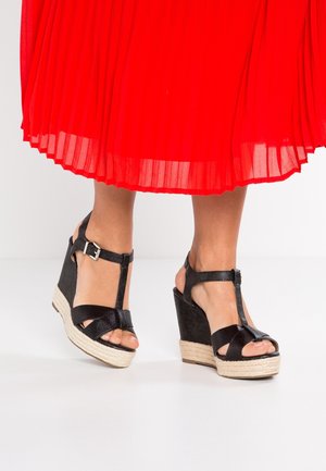 Piernas con sandalias negras de cuña con tiras en el tobillo y una falda midi plisada roja, de pie sobre un suelo blanco con un fondo claro.