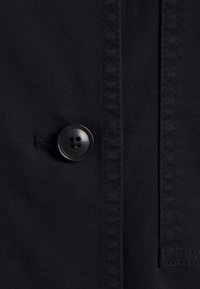 Zwarte stof close-up met een viergaatjesknop en gestructureerde stiksels. Gladde textuur met een lichte glans en zichtbare zakdetails.