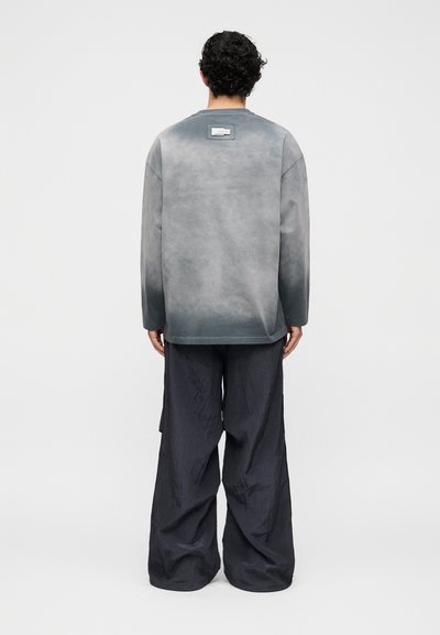 Άνδρας με σγουρά μαλλιά, ντυμένος με γκρι μακρυμάνικη μπλούζα με μικρή ετικέτα στο πάνω μέρος της πλάτης και φαρδιά μαύρη παντελόνια, που στέκεται με την πλάτη γυρισμένη.