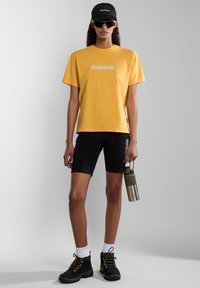 Geel katoenen t-shirt met logo-print, zwarte shorts, zwarte laarzen met gele veters, een groene waterfles vasthoudend. Witte sokken en een zonnebril.