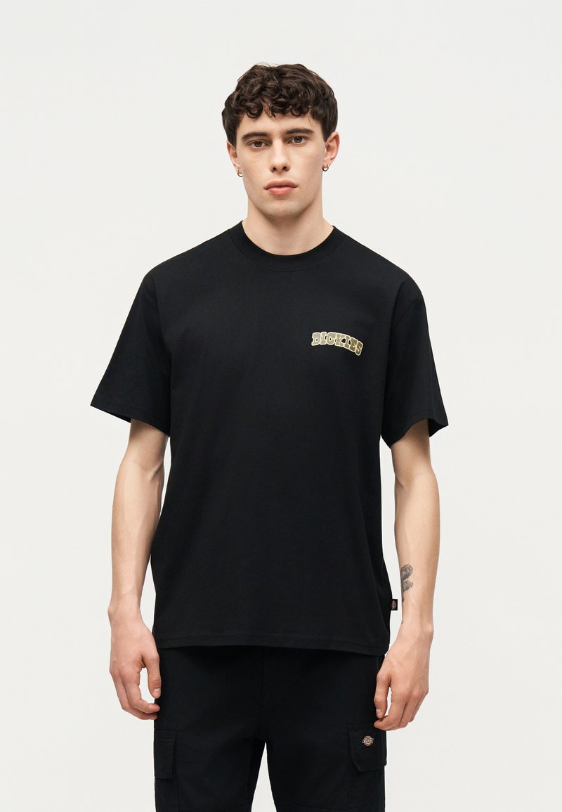 Giovane uomo con capelli corti e ricci, indossa una t-shirt nera Dickies e pantaloni neri abbinati, in piedi davanti a uno sfondo bianco uniforme.