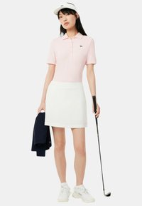 Polo in morbido tessuto rosa, gonna bianca e scarpe da ginnastica bianche. Tiene un bastone da golf, indossa un visiera bianca e ha un giubbotto blu marino drappeggiato.