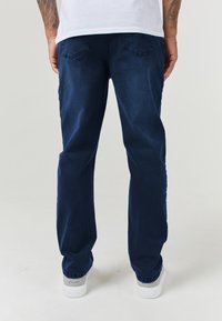 Jeans de mezclilla azul marino con corte recto, bolsillos traseros visibles y una ligera textura. Combinados con zapatillas blancas en la parte inferior.