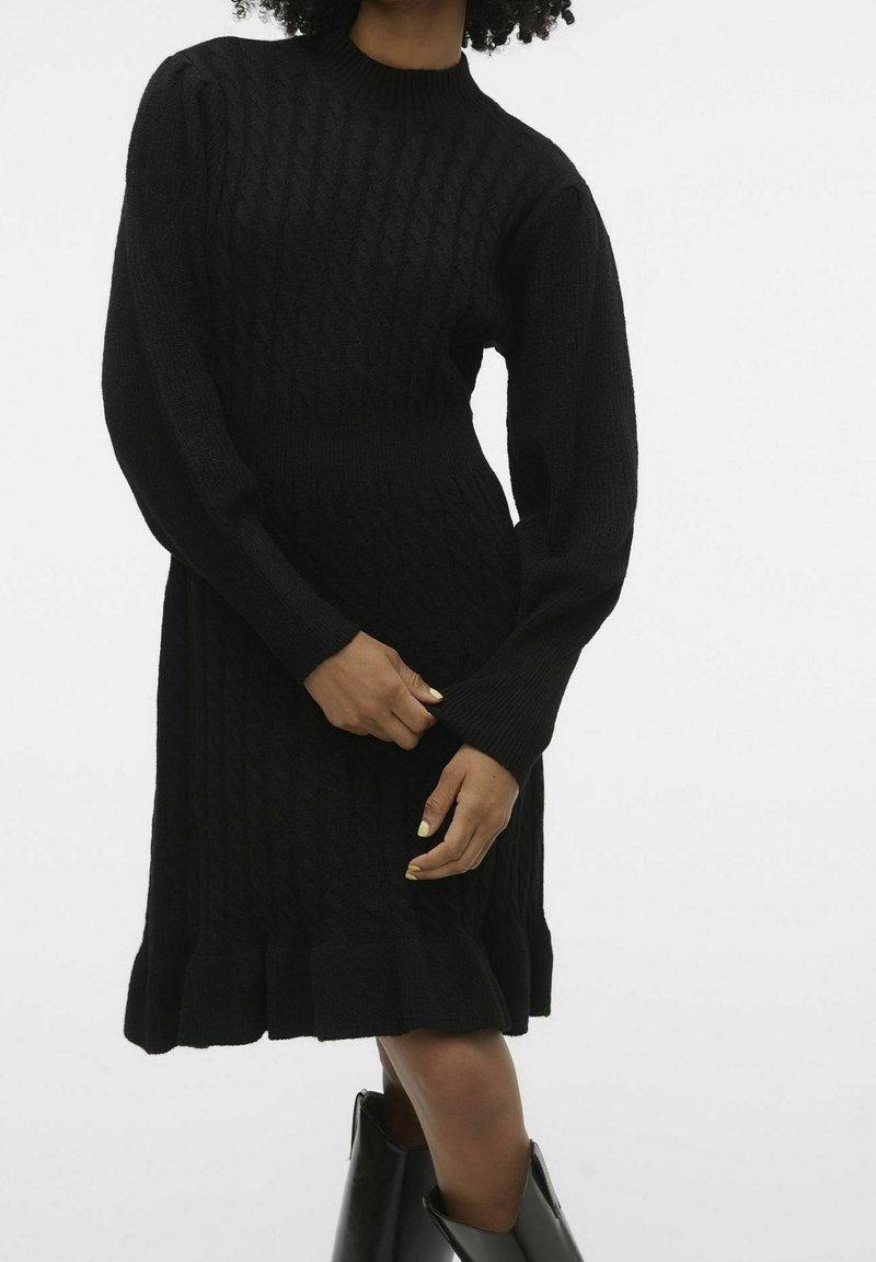 Robe noire en tricot avec un motif en torsades, un col à côtes, des manches longues et un ourlet volanté. La texture semble douce et confortable.