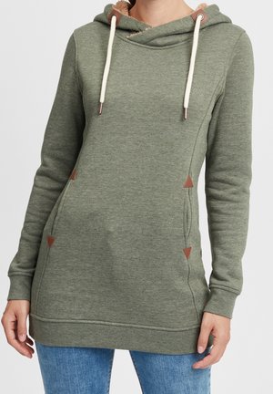 Frau trägt olivgrünen Kapuzenpullover mit weißen Kordeln, Kängurutaschen und braunen dreieckigen Akzenten, kombiniert mit blauer Jeans.