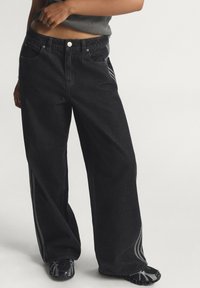 Μαύρο τζιν με φαρδιές πόδες, υλικό ντένιμ, σχέδιο με πέντε τσέπες, ασημένιο κουμπί στο κλείσιμο, ρίγες στο πλάι με αντίθετα χρώματα, συνδυασμένο με μαύρα παπούτσια.