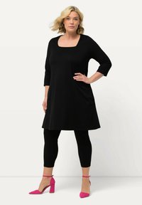 Schwarzes knielanges Kleid mit quadratischem Ausschnitt und 3/4-Ärmeln, kombiniert mit schwarzen Leggings und leuchtend rosa High Heels.