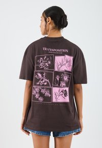 Brun oversized t-skjorte med rosa blomsterillustrasjoner og tekst på ryggen, med en avslappet passform og korte ermer.