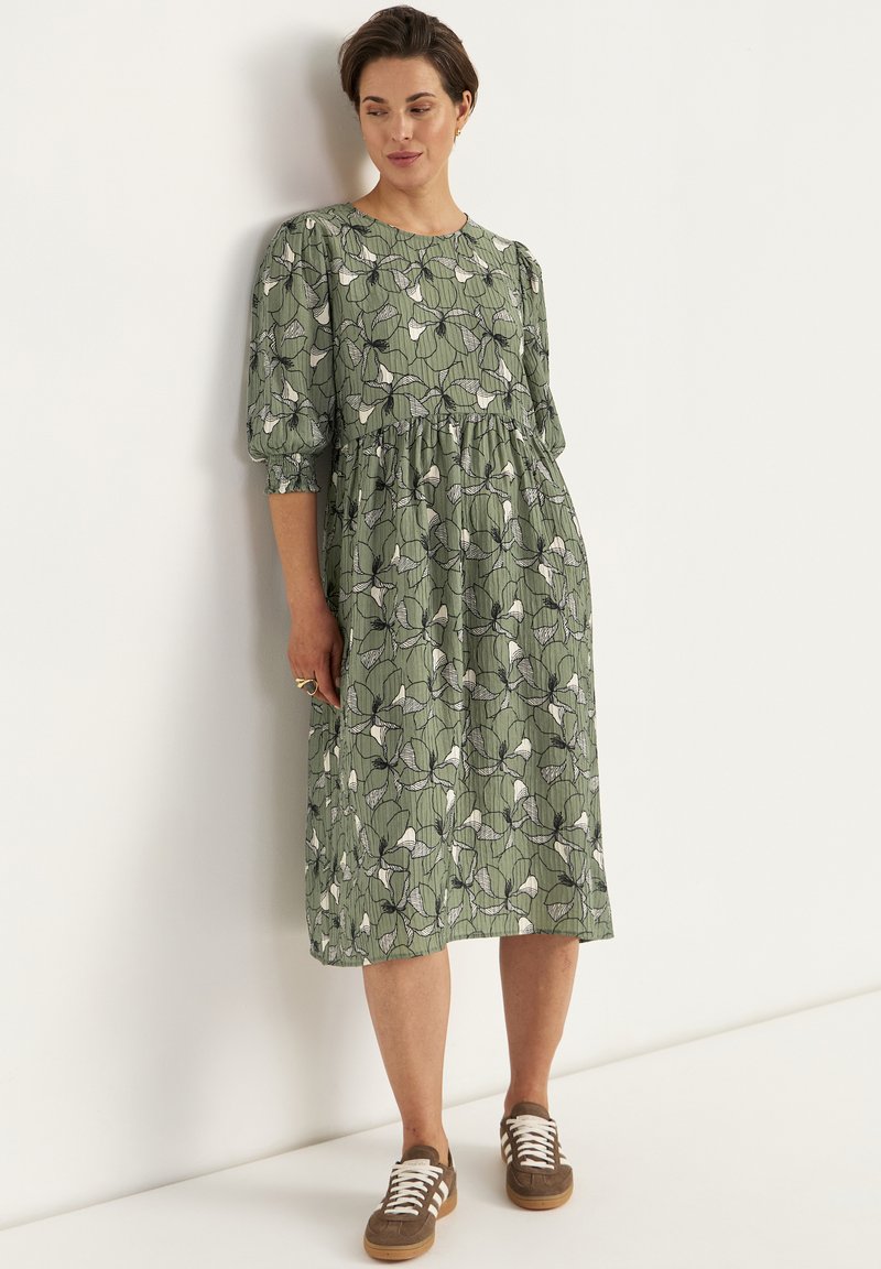 Grøn kjole med blomsterprint, der har pufærmer og en indsnævret talje. Kombineret med brune sneakers og en ring på venstre hånd.