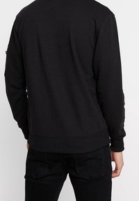 Persona vistiendo un sudadera negra de manga larga sencilla y jeans negros, vista desde atrás contra un fondo claro.